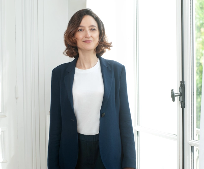Maître Raphaële Secnazi-Leiba debout dans un intérieur lumineux, son bureau, vêtue d’un blazer bleu marine, photo professionnelle prise à Paris